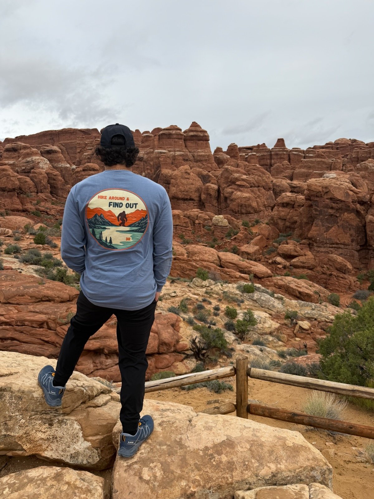 A person in a black cap and blue jacket with a hiking graphic on the back stands on rocks, overlooking a red rock canyon landscape with large formations and sparse vegetation under an overcast sky.