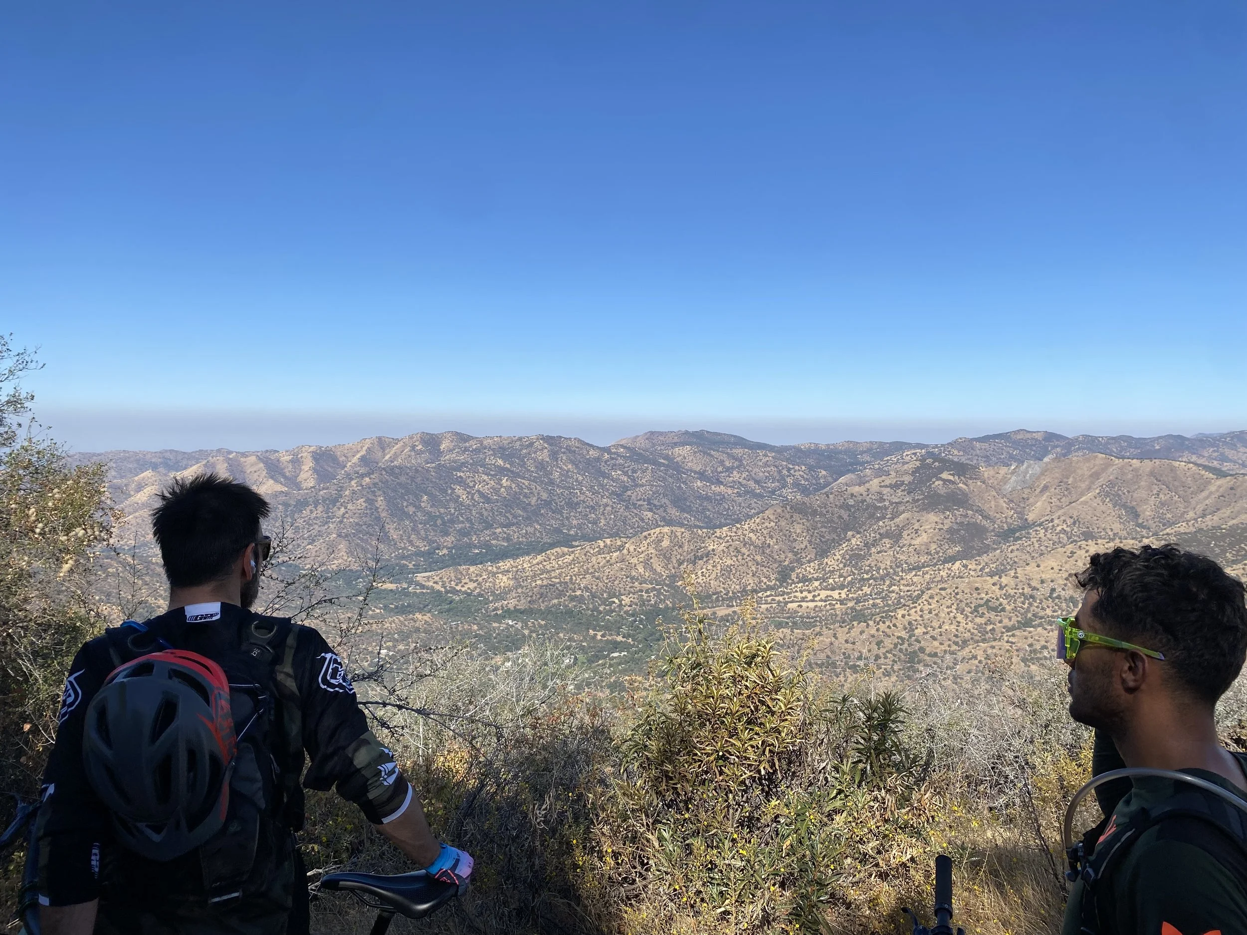 Two men with bike helmets in the foreground overlooking a mountain range with dry, rugged hills and sparse vegetation, under a clear blue sky.