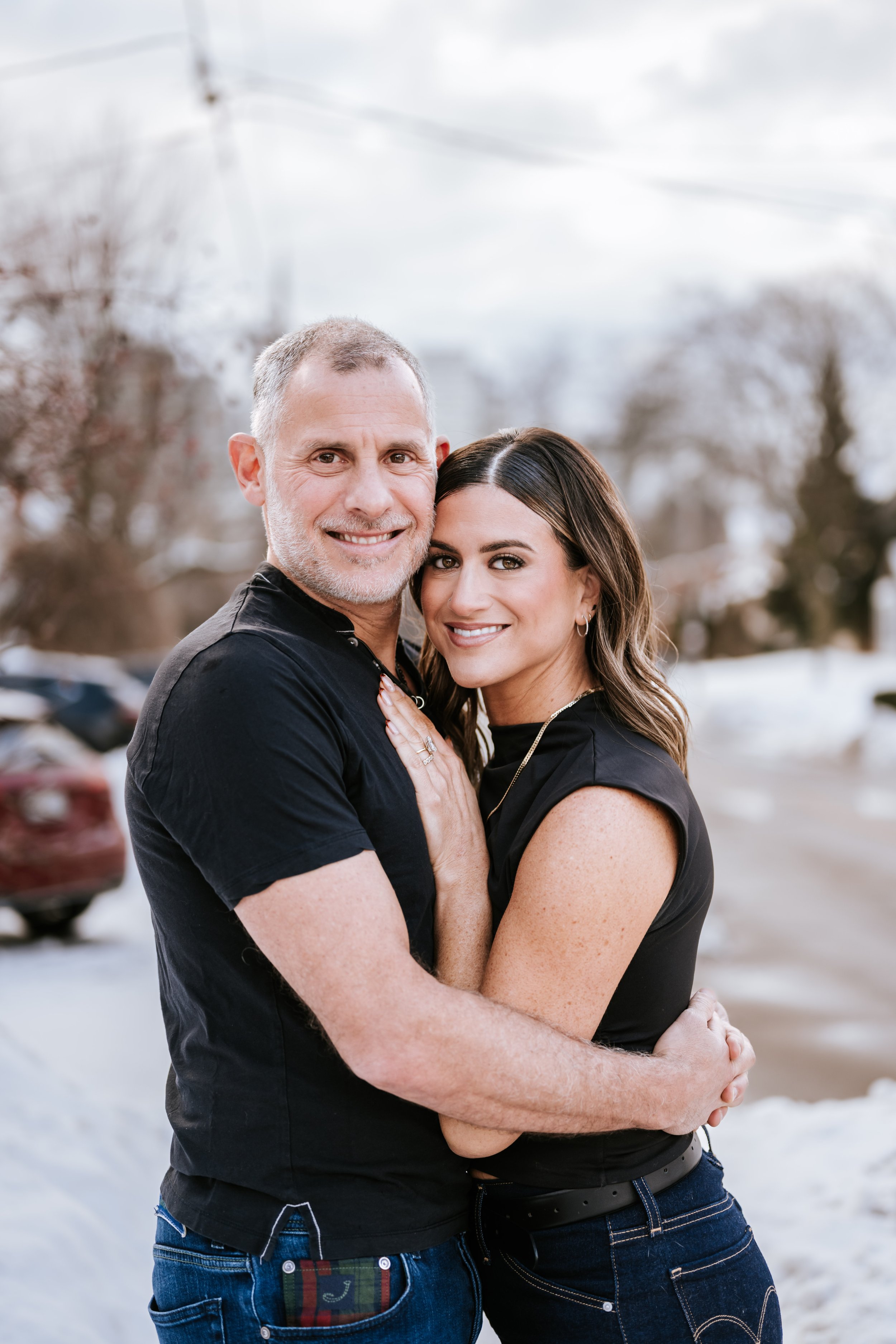 A middle-aged man and a young woman are standing outdoors, close together, smiling at the camera. The man has short gray hair, a beard, and is wearing a black shirt. The woman has long brown hair, light makeup, and is wearing a black top. They are embracing with the man's arms around the woman's shoulders and the woman's hands on the man's chest. The background shows trees and a cloudy sky, with a blurred view of cars and snow on the ground.