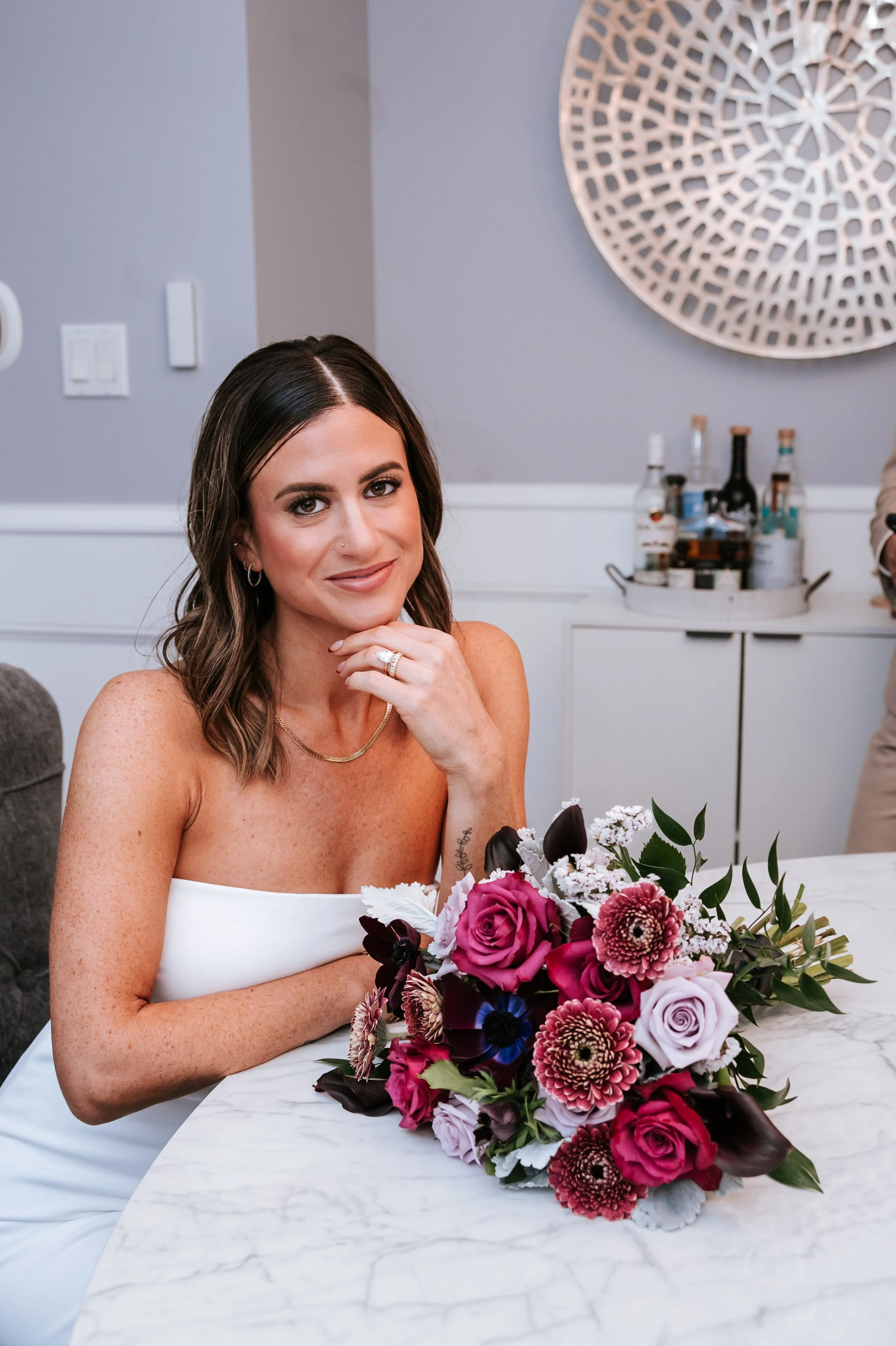 A woman with dark brown hair, light skin and a small nose piercing, wearing a white strapless dress, seated at a marble table with a bouquet of pink, purple, and burgundy flowers in front of her.