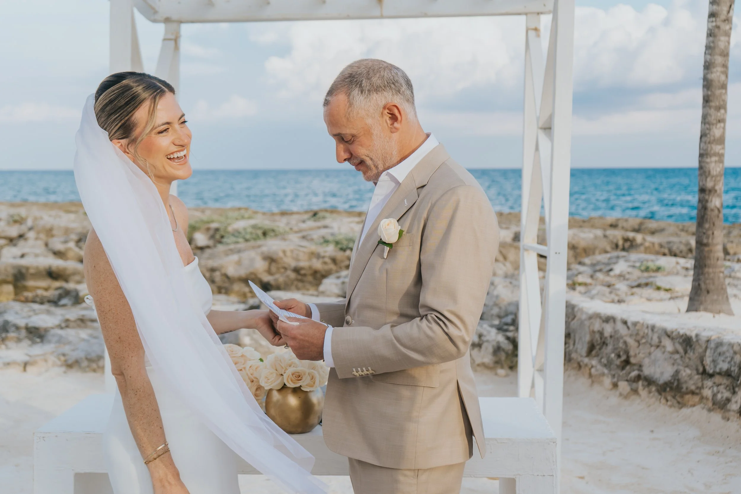 A bride in a white wedding dress and veil smiling as she looks at an officiant during a beach wedding ceremony, with the ocean in the background and a white arch.