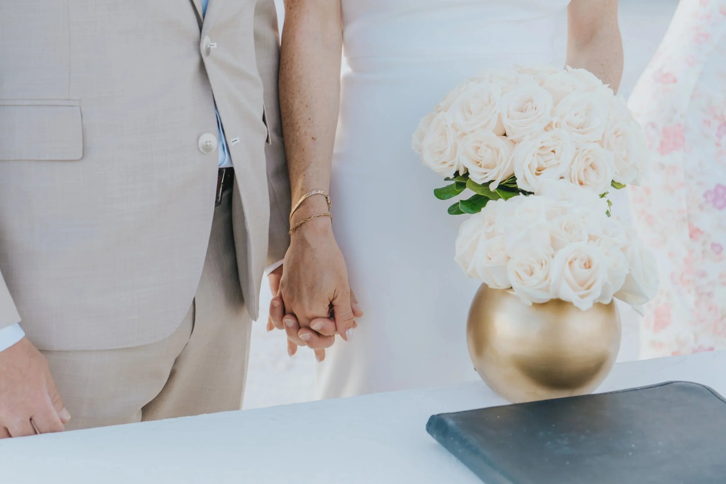 Close-up of a couple holding hands at a wedding ceremony, with a bouquet of cream-colored roses in a gold vase nearby.