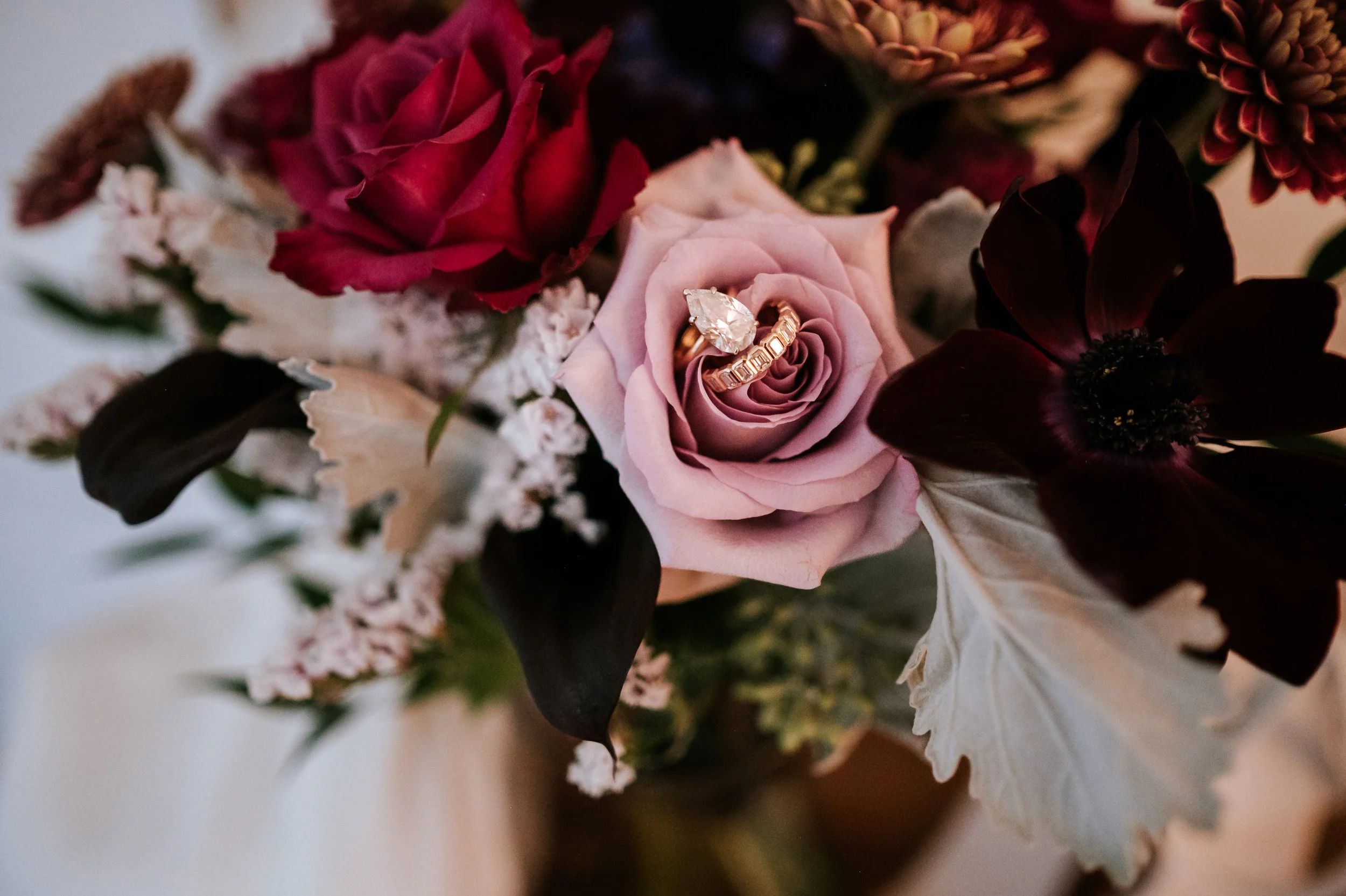 Close-up of a bouquet with pink and dark red roses, white and beige flowers, and a gold ring with a large central gem placed on a pink rose.