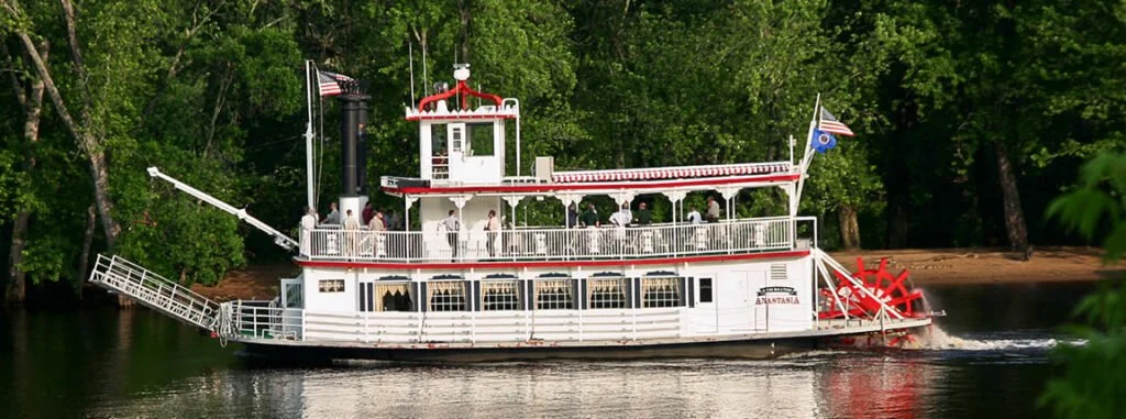 A paddlewheel riverboat named 'Anastasia' on a river, surrounded by green trees with people onboard.