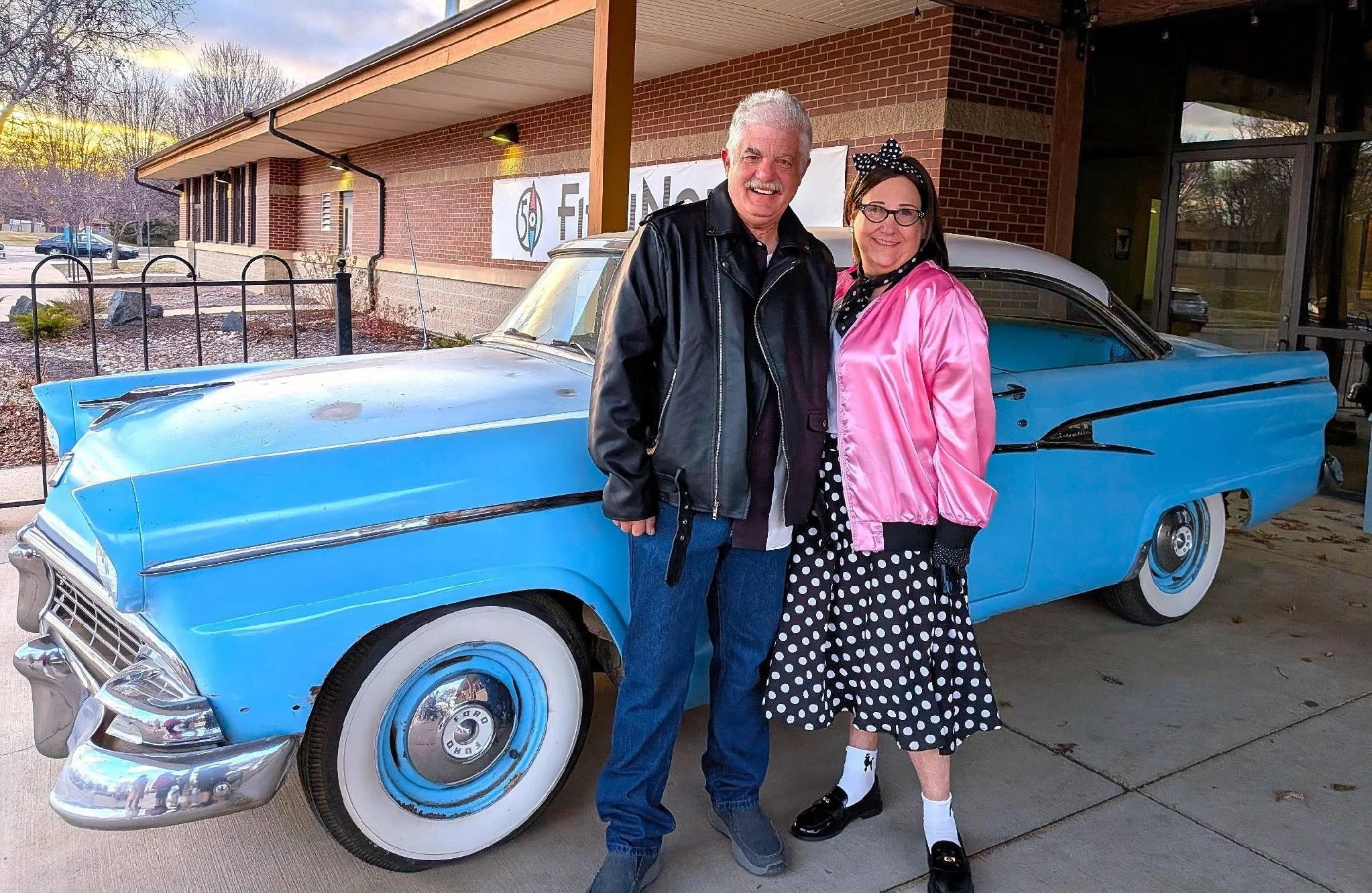 A smiling man and woman stand together outside next to a vintage blue car with a white roof, in front of a brick building with large windows and a sign that reads 'FINDING' and some other text. The man wears a black leather jacket and jeans, and the woman is dressed in a pink satin jacket, a polka dot dress, and a headband with polka dots.