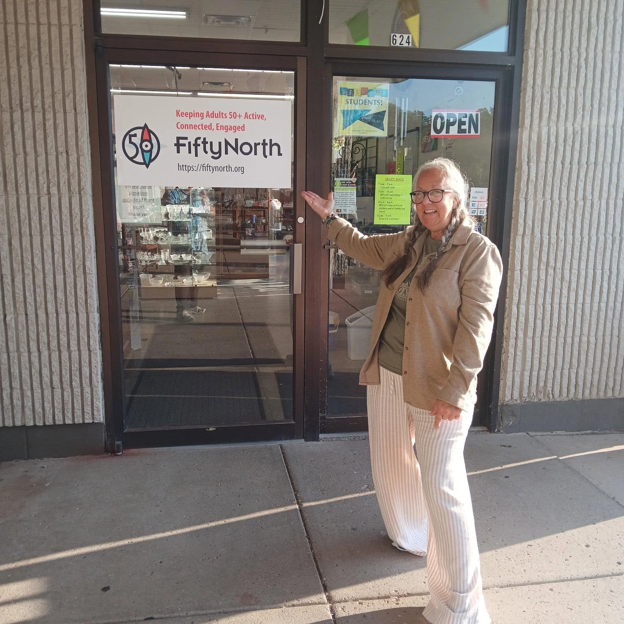 A woman standing outside a building with glass doors, pointing at a sign that reads 'Keeping Adults 50+ Active, Connected, Engaged,' with a logo for FiftyNorth and a website URL. The woman has gray curly hair, glasses, and is wearing a beige jacket and striped pants. The building has a brick exterior, and there are various signs and posters in the windows.