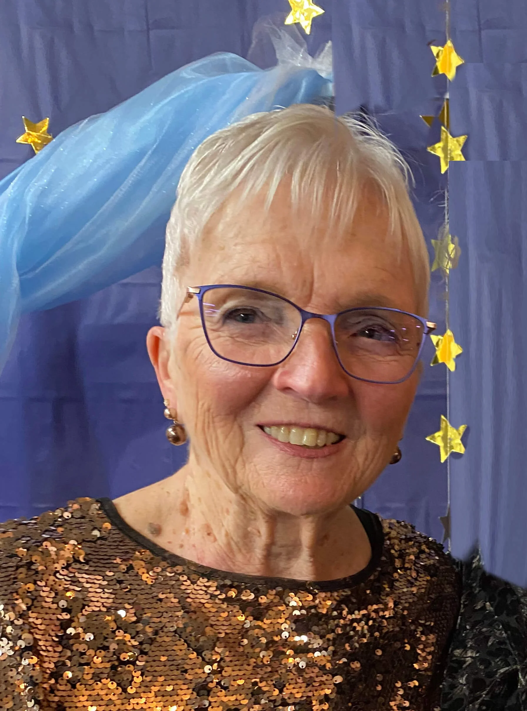 Smiling older woman with glasses, short gray hair, wearing a black sequined top, standing in front of a blue backdrop with hanging gold star decorations.