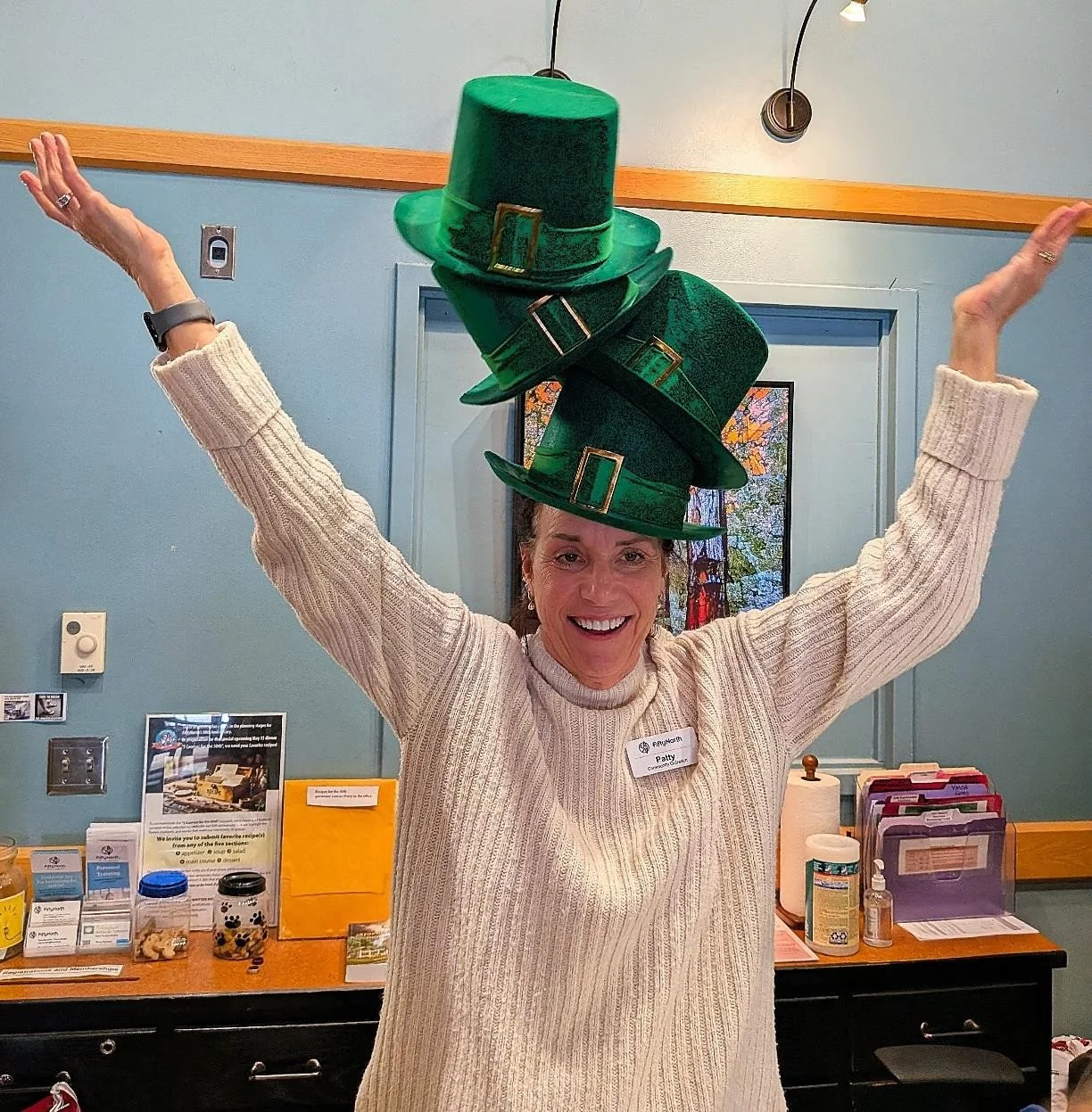 Woman smiling and wearing three green St. Patrick's Day hats stacked on her head with arms raised in celebration.