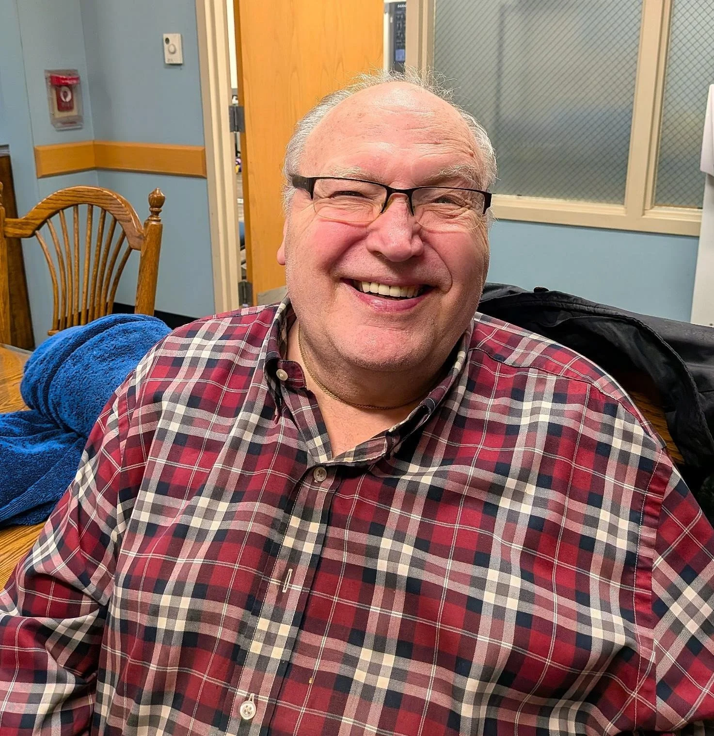 A smiling middle-aged man with glasses and a plaid shirt sitting at a table in a room with light blue walls, wooden chairs, and a window with a wire mesh.