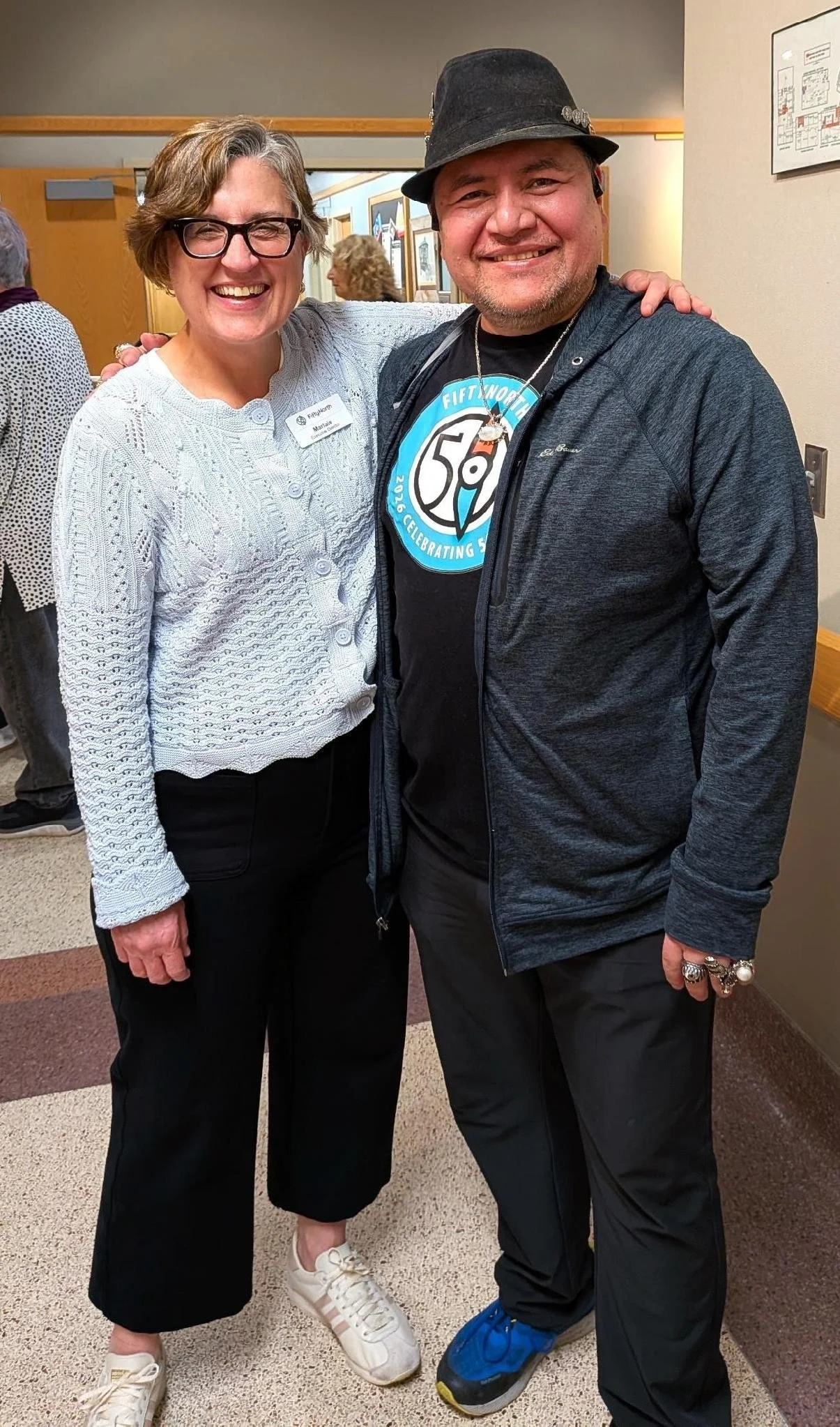 Two smiling people standing close together in an indoor setting, with their arms around each other. The woman on the left has short gray hair, wears glasses, a white knit sweater, black pants, and white sneakers. The man on the right has a beard, wears a black hat, a black T-shirt with a blue and white logo, a gray zip-up jacket, black pants, and blue shoes.