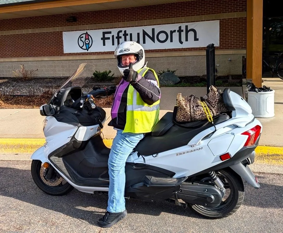 A person on a white touring scooter, wearing a helmet and a reflective safety vest, sitting and giving a thumbs-up in front of a building with a Fifty North sign, with leopard print bags on the back of the scooter.