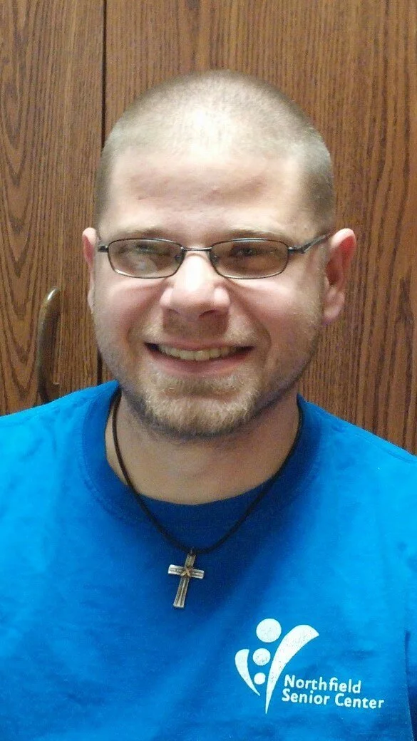 A smiling man with glasses and a short beard wearing a blue shirt with the Northfield Senior Center logo, a cross necklace, and standing in front of a wooden door.