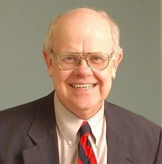 A smiling elderly man with glasses, wearing a dark suit, a light-colored shirt, and a striped red and blue tie.