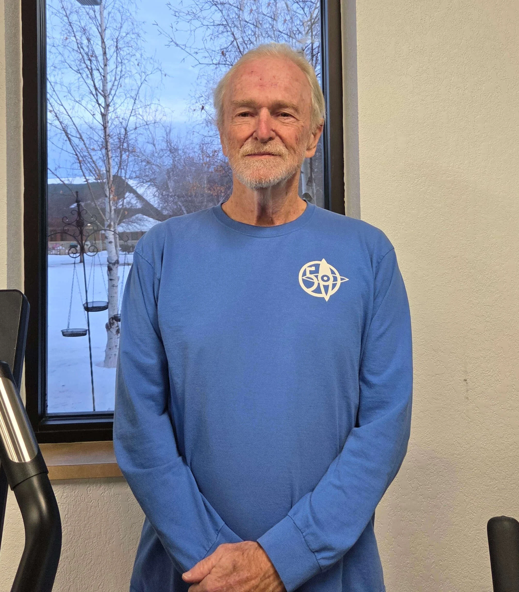 Older man with white hair, beard, wearing a blue long sleeve shirt, standing indoors in front of a window with leafless trees outside, and a wall with textured paint.
