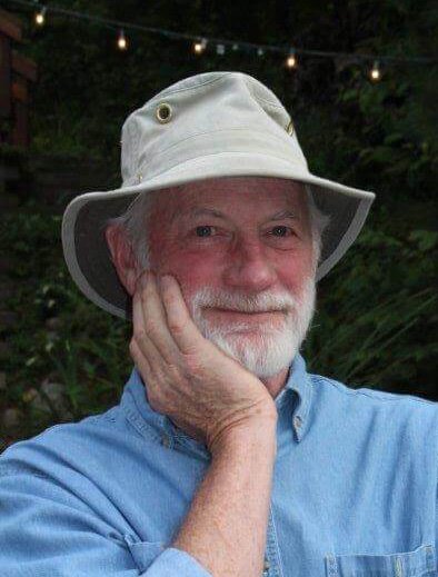 An older man with a white beard and a hat, smiling and resting his face on his hand outdoors with string lights and foliage in the background.