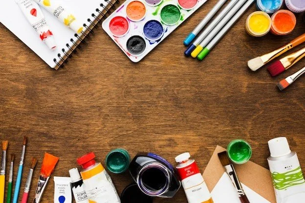 Art supplies including watercolor paints, brushes, markers, and notebooks arranged on a wooden surface.