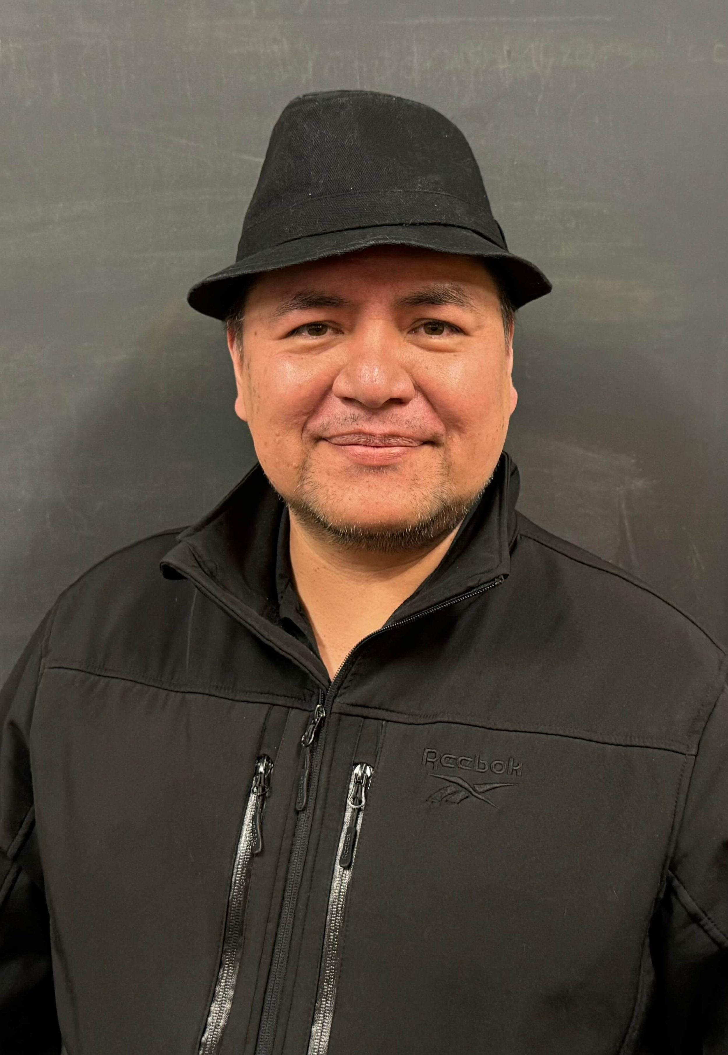 A man with short dark hair and light brown skin smiling, wearing a black bucket hat and a black Reebok jacket, standing against a plain dark wall.