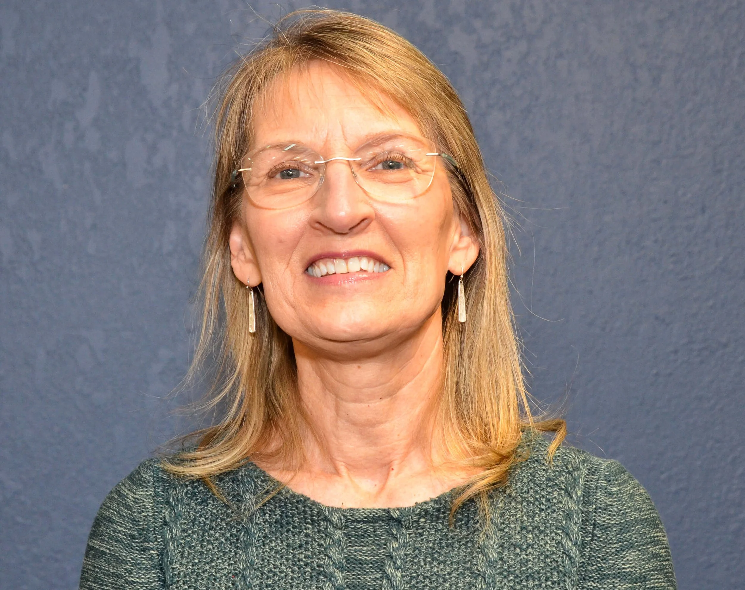 Close-up of a smiling middle-aged woman with glasses, earrings, and shoulder-length blond hair, posed against a plain gray background.