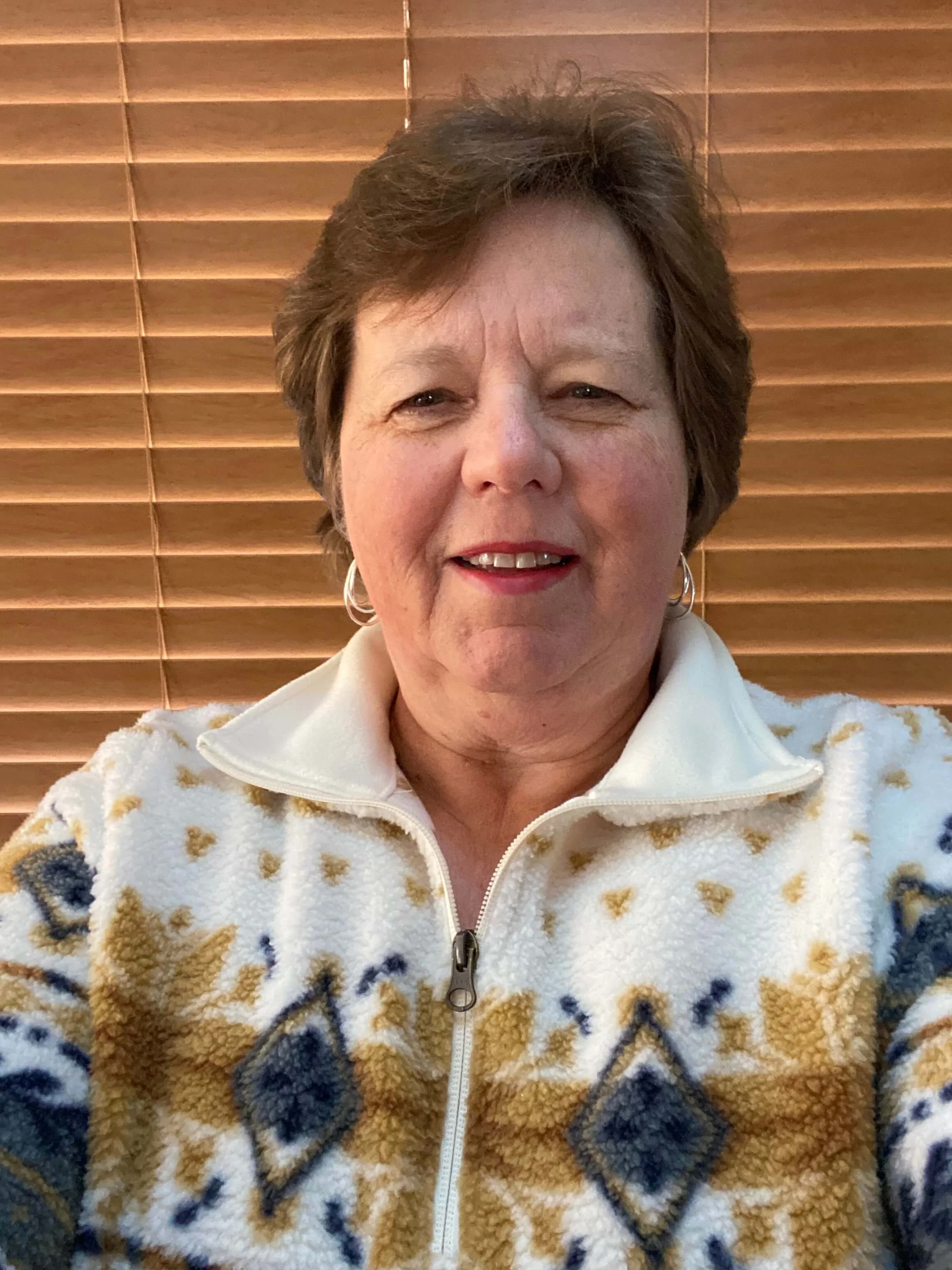 A woman with short brown hair wearing a white sherpa jacket with a zip, patterned with brown, black, and white. She is smiling slightly and standing in front of closed wooden blinds.