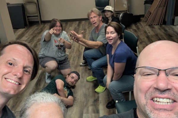 Group of eight people gathered in a room, some sitting and some lying on the floor, smiling and taking a selfie.