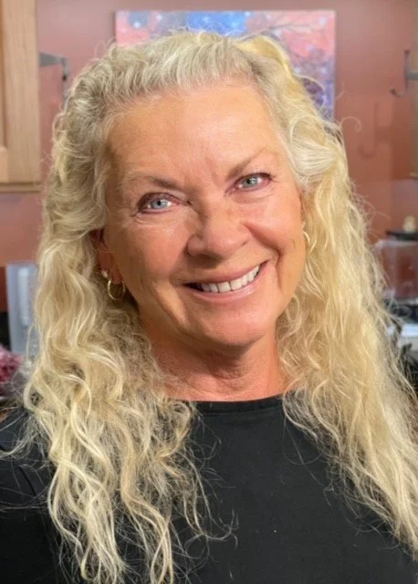 A smiling elderly woman with long, curly blonde hair, wearing a black top and small earrings, standing indoors with a blurred background.