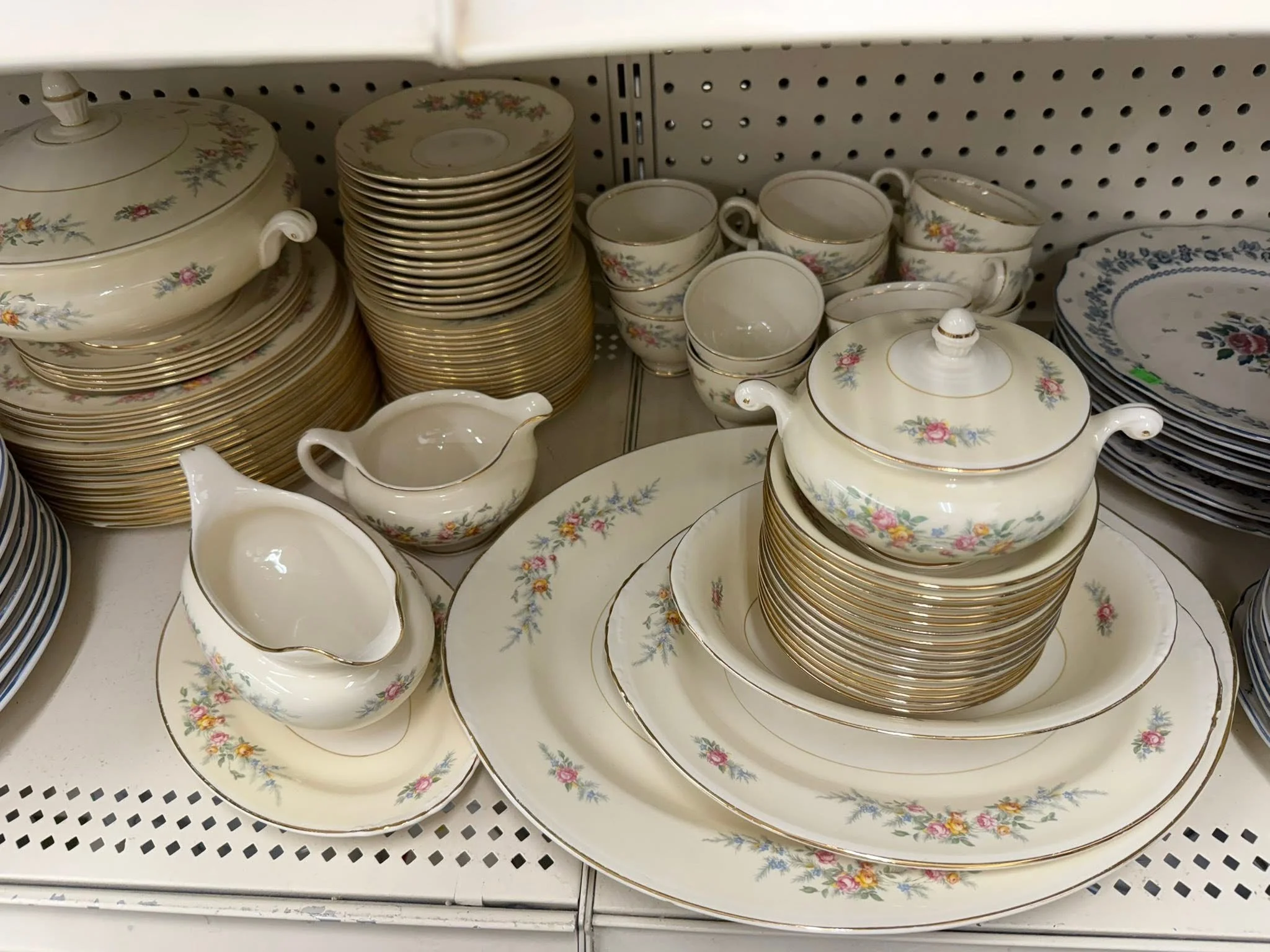 Stacked floral-patterned china dishes on a store shelf, including teacups, saucers, plates, and covered serving dishes with gold trim.