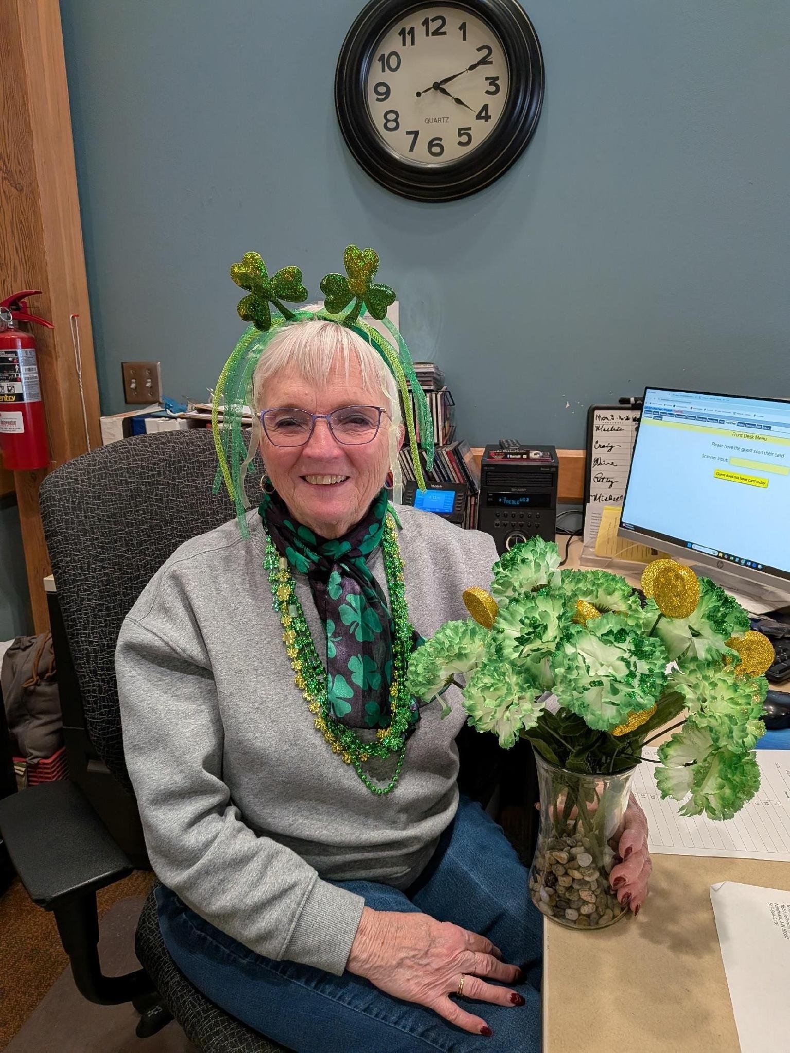 An elderly woman with white hair and glasses, smiling, decorated with green shamrock headbands and necklaces, holding a large St. Patrick's Day floral arrangement in a glass vase. She is sitting at a desk in an office with a computer, books, and a whiteboard behind her. The clock on the wall shows 2:10.