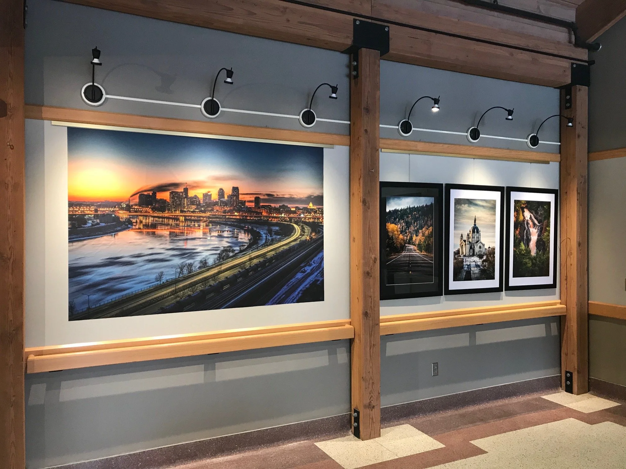 Photograph of a gallery wall displaying framed photos of cityscapes and landscapes, with a colorful sunset over a city skyline, mounted on a white wall with wooden beams and track lighting above.