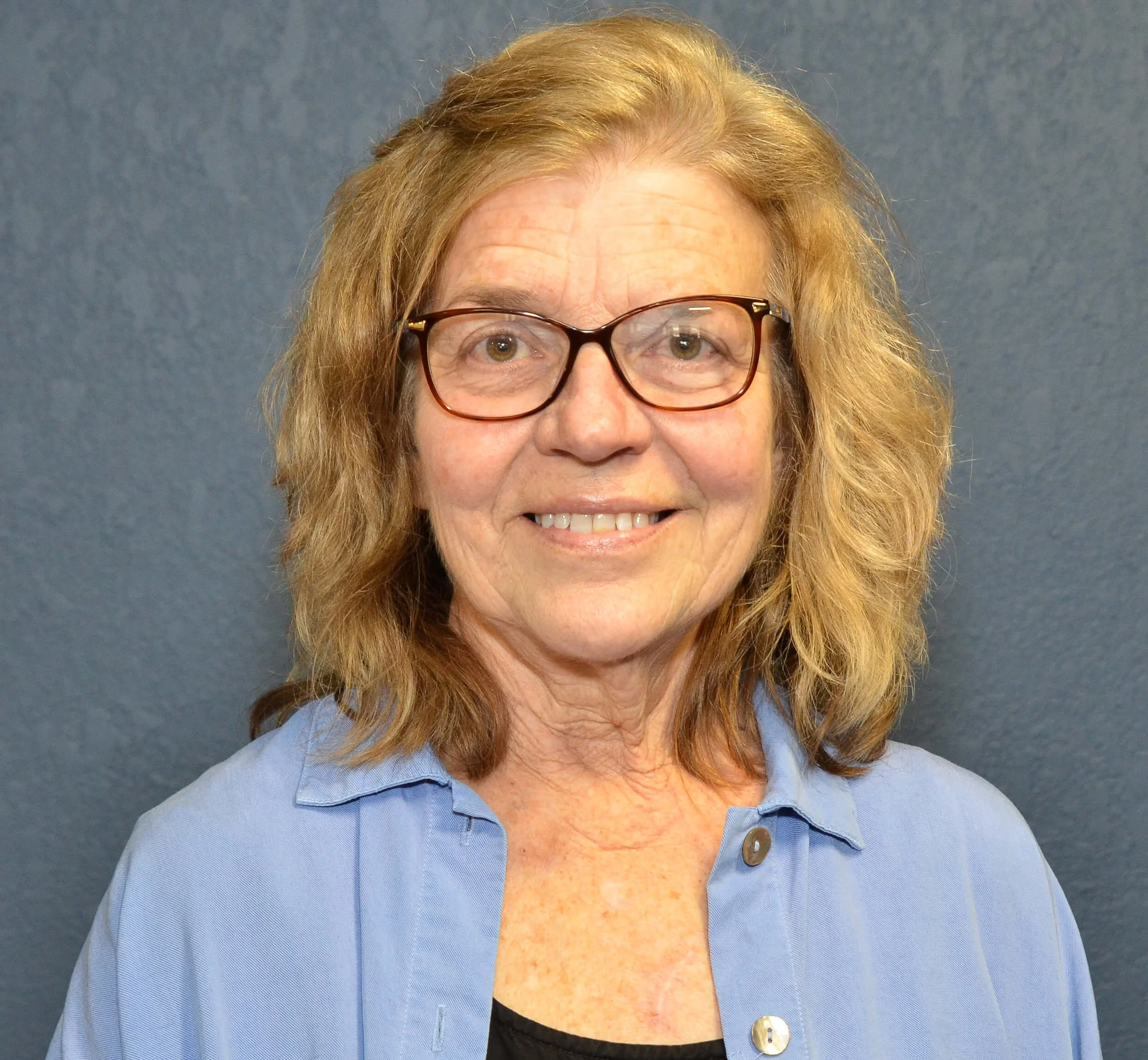 Close-up of a middle-aged woman with blonde hair, glasses, wearing a blue shirt, smiling, against a gray background.