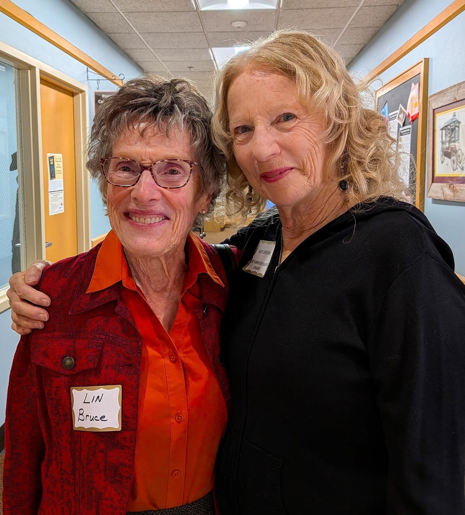 Two women smiling and hugging in an indoor setting with light blue walls and framed artwork. One woman has short gray hair and glasses, wearing a red jacket and orange shirt with a name tag reading 'LIN Bruce'. The other woman has curly blonde hair, wearing a black top and earrings, with a name tag on her chest.