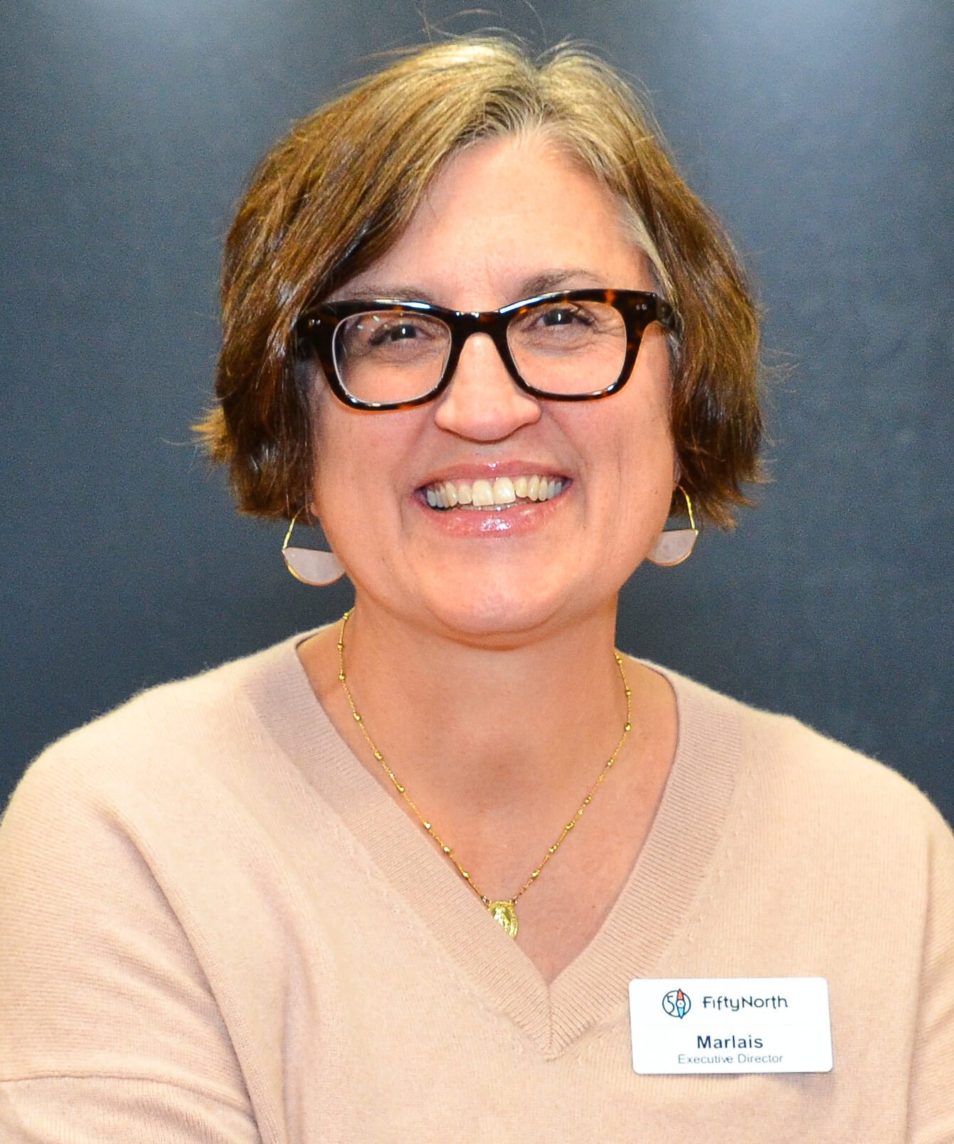 A woman with short, light brown hair and glasses smiling, wearing a beige sweater and gold jewelry, with a name tag that reads Marlais, Executive Director, at FidNorth.