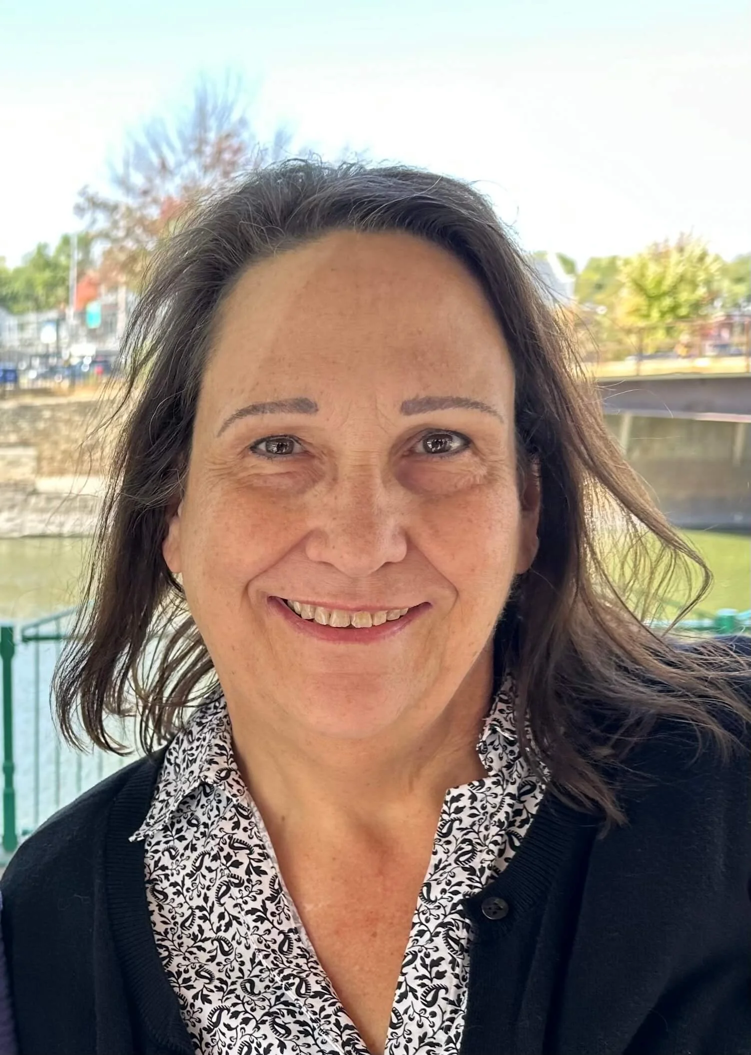 A middle-aged woman with shoulder-length dark hair and light skin, smiling and wearing a black cardigan over a white blouse with a black leaf pattern, standing outdoors with a blurred background of trees and a body of water.