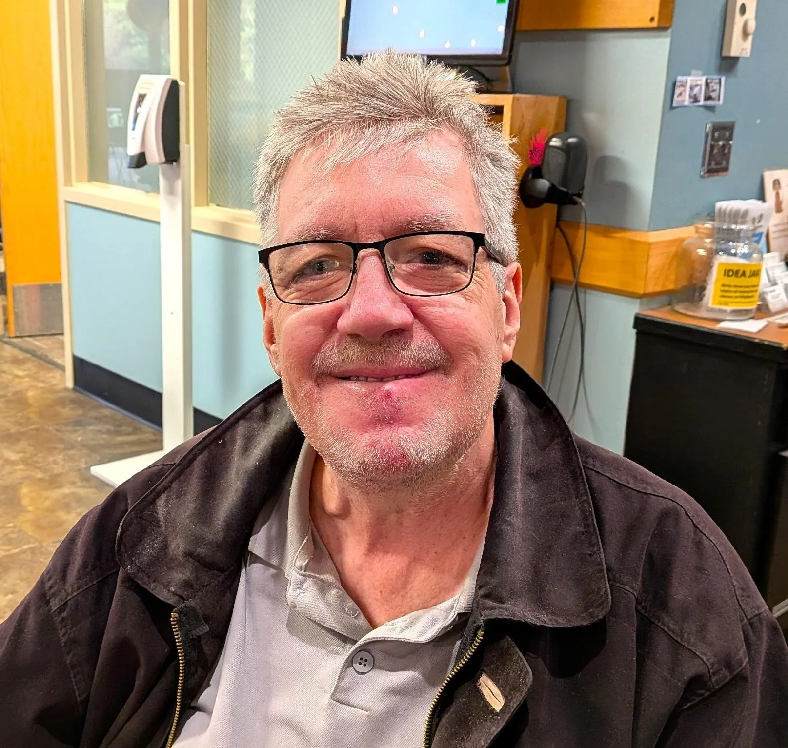 A smiling man with glasses and gray hair sitting in a room with a computer monitor, bottles, and various items behind him.