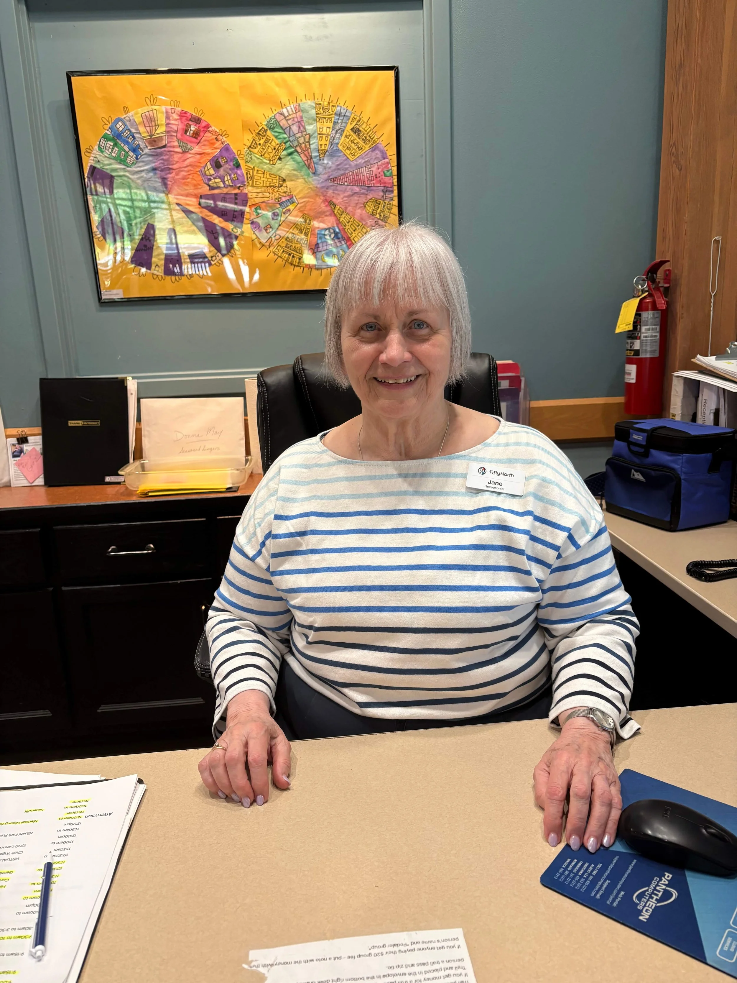 An elderly woman with short gray hair, wearing a striped shirt, smiling while sitting at a desk with papers, a pen, and a computer mouse in front of her. Behind her is a colorful abstract art piece on the wall, and to her right is a red fire extinguisher.