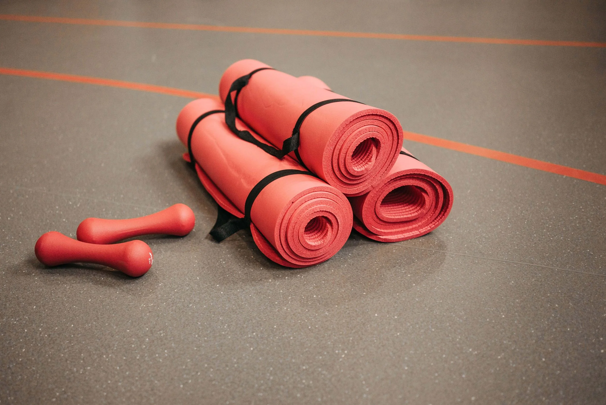 Four pink yoga mats rolled up and secured with black straps, with two pink dumbbells nearby, on a gym floor with orange lines.