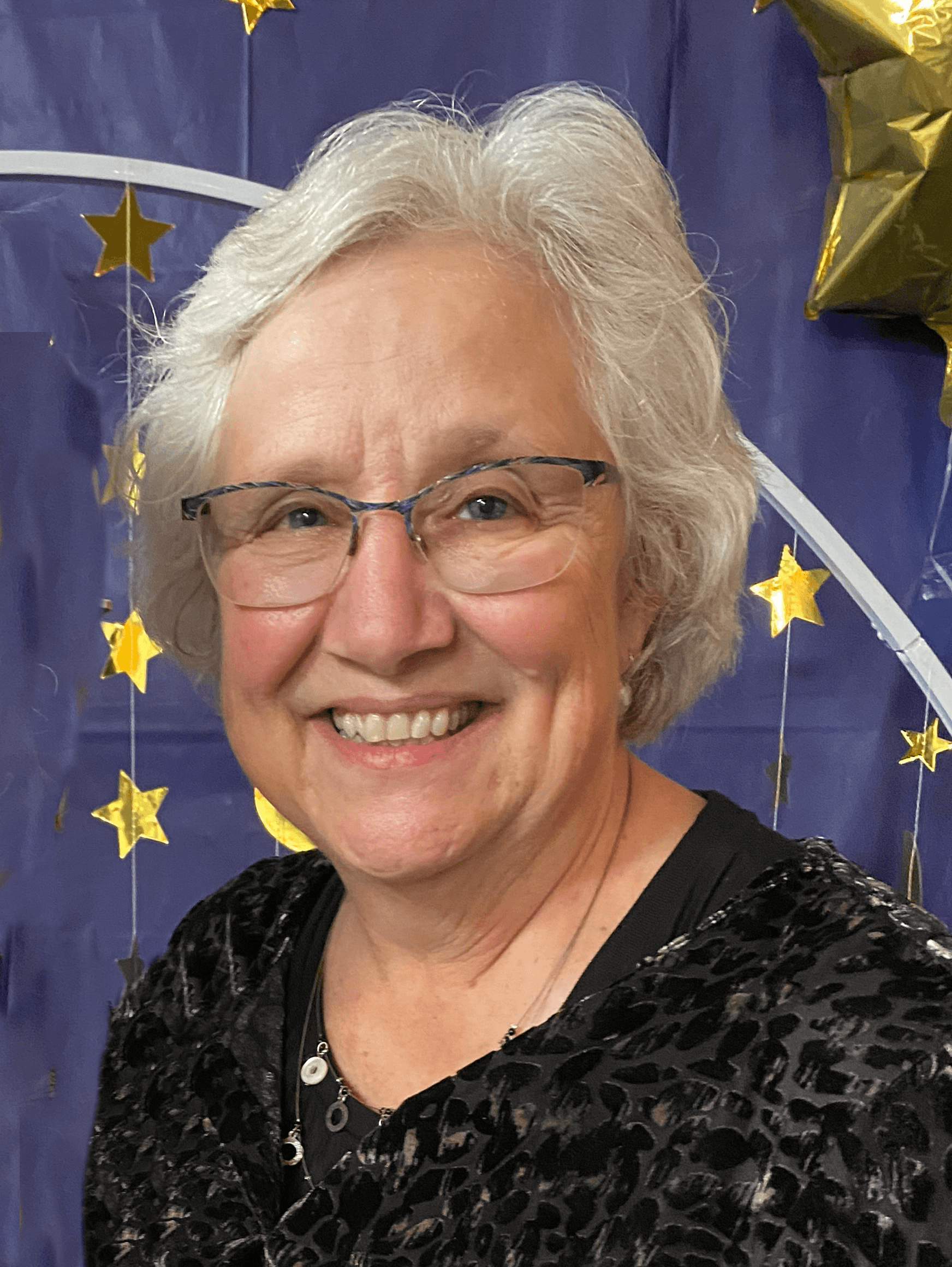 Smiling elderly woman with glasses, short gray hair, and black patterned top, standing in front of a blue backdrop decorated with gold star-shaped balloons and a white arch with stars.