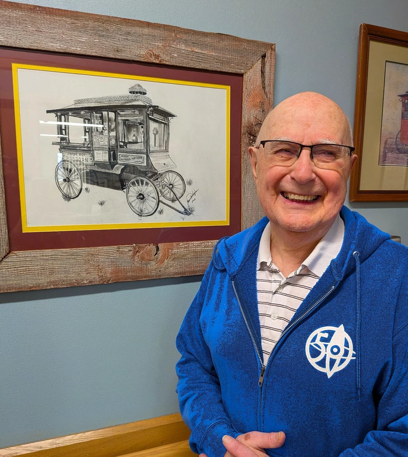 A smiling man with glasses wearing a blue jacket and striped polo shirt, standing next to framed artwork of a vintage popcorn cart on a wooden wall.