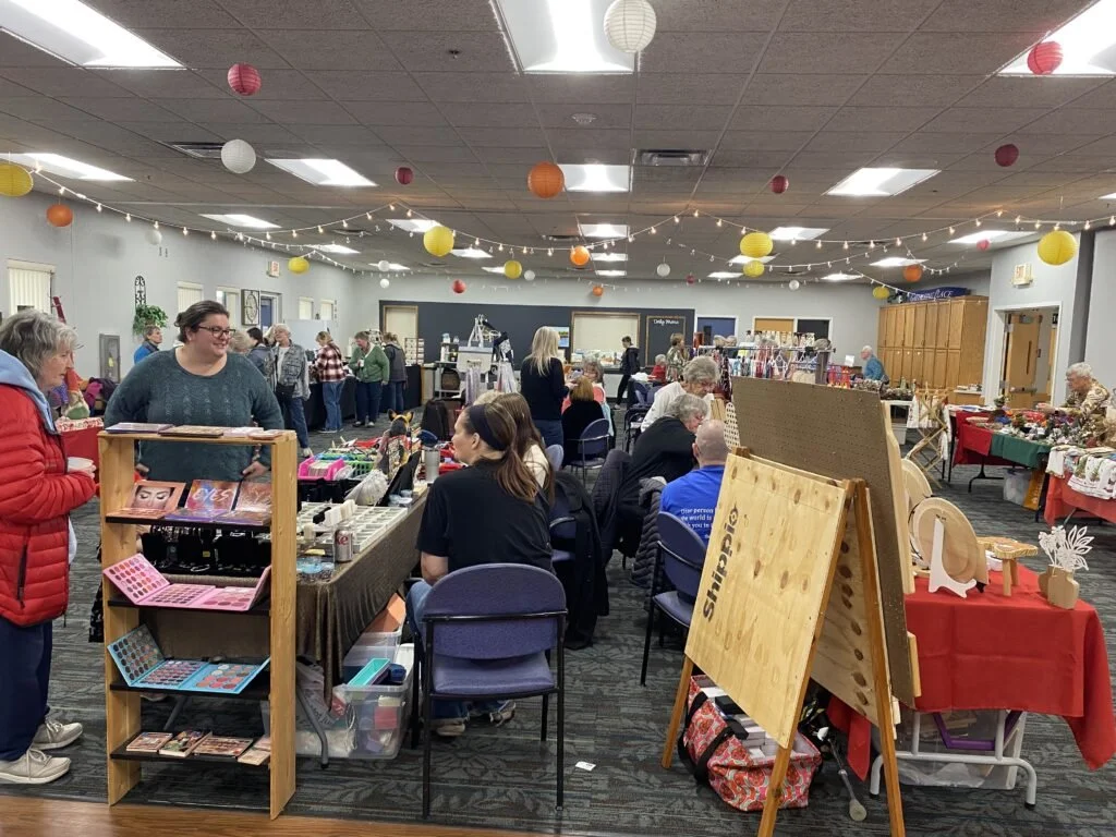 People browsing and shopping at a craft fair or indoor market with decorated ceiling and tables displaying handmade items.