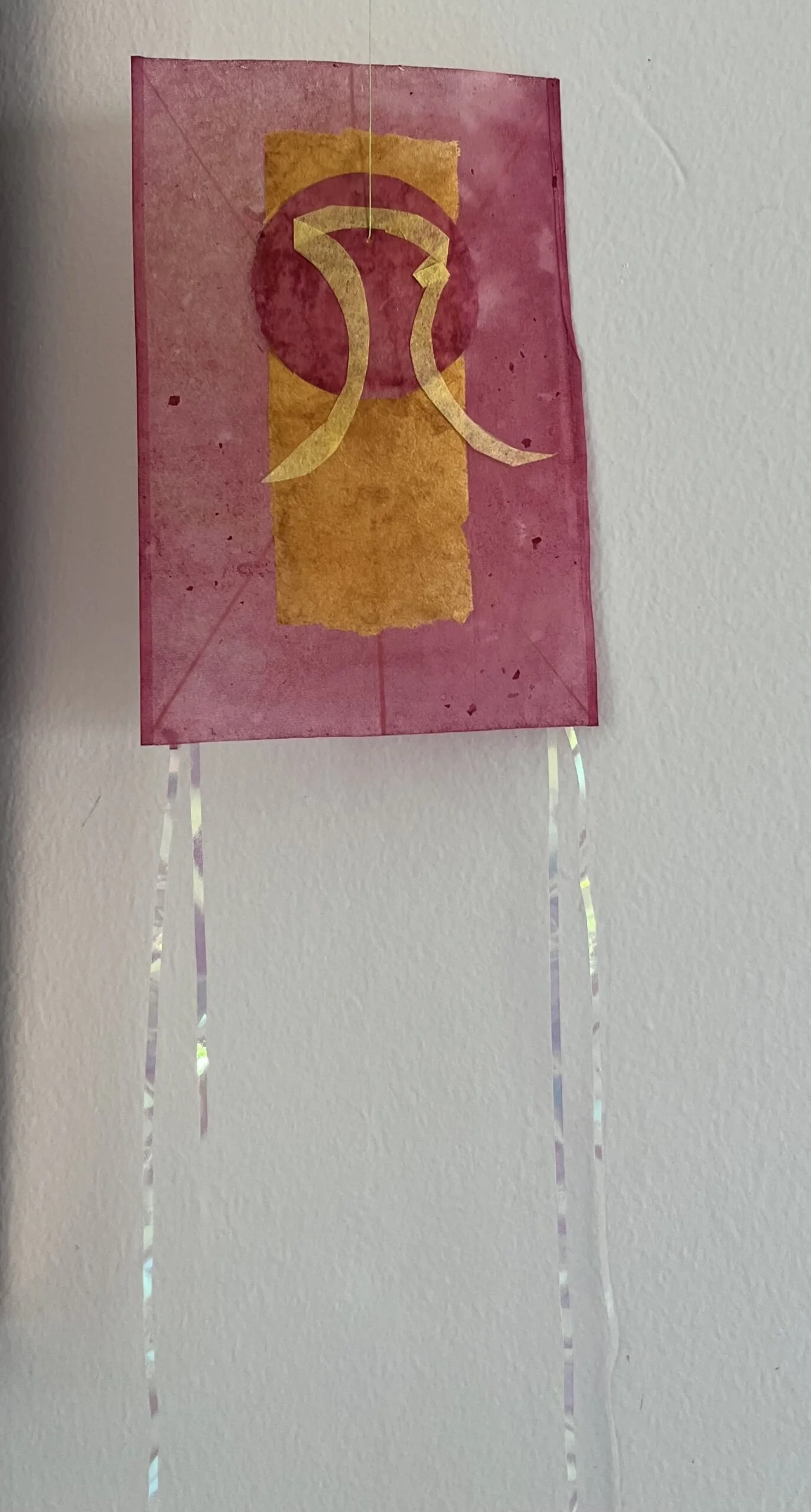 Pink paper lantern decorated with yellow and pink designs, hanging on a white wall with shiny metallic streamers.