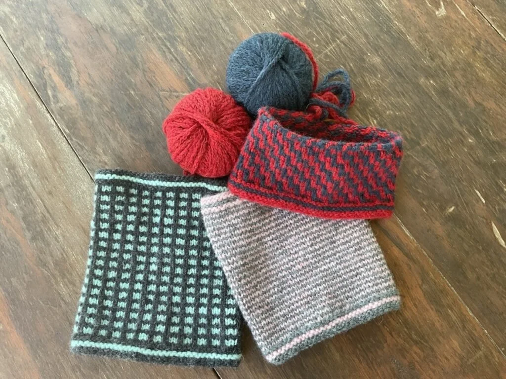 Two balls of yarn, one red and one dark blue, with a red and black patterned knit hat and a striped gray scarf on a wooden surface.