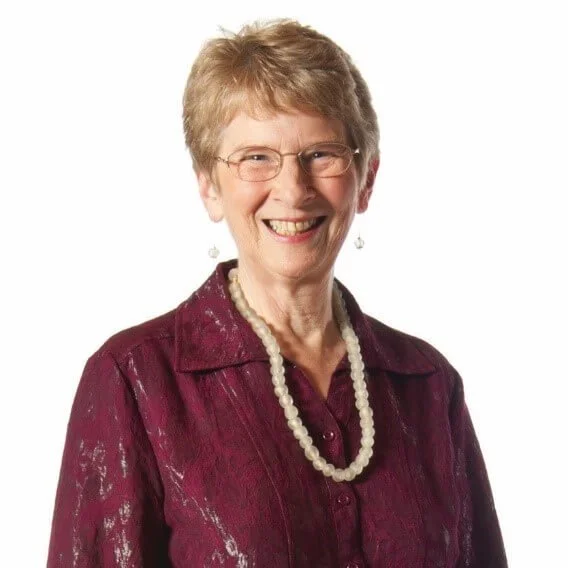 A smiling elderly woman with short, light-colored hair, wearing glasses, a pearl necklace, earrings, and a dark maroon blouse with a textured pattern, standing against a white background.