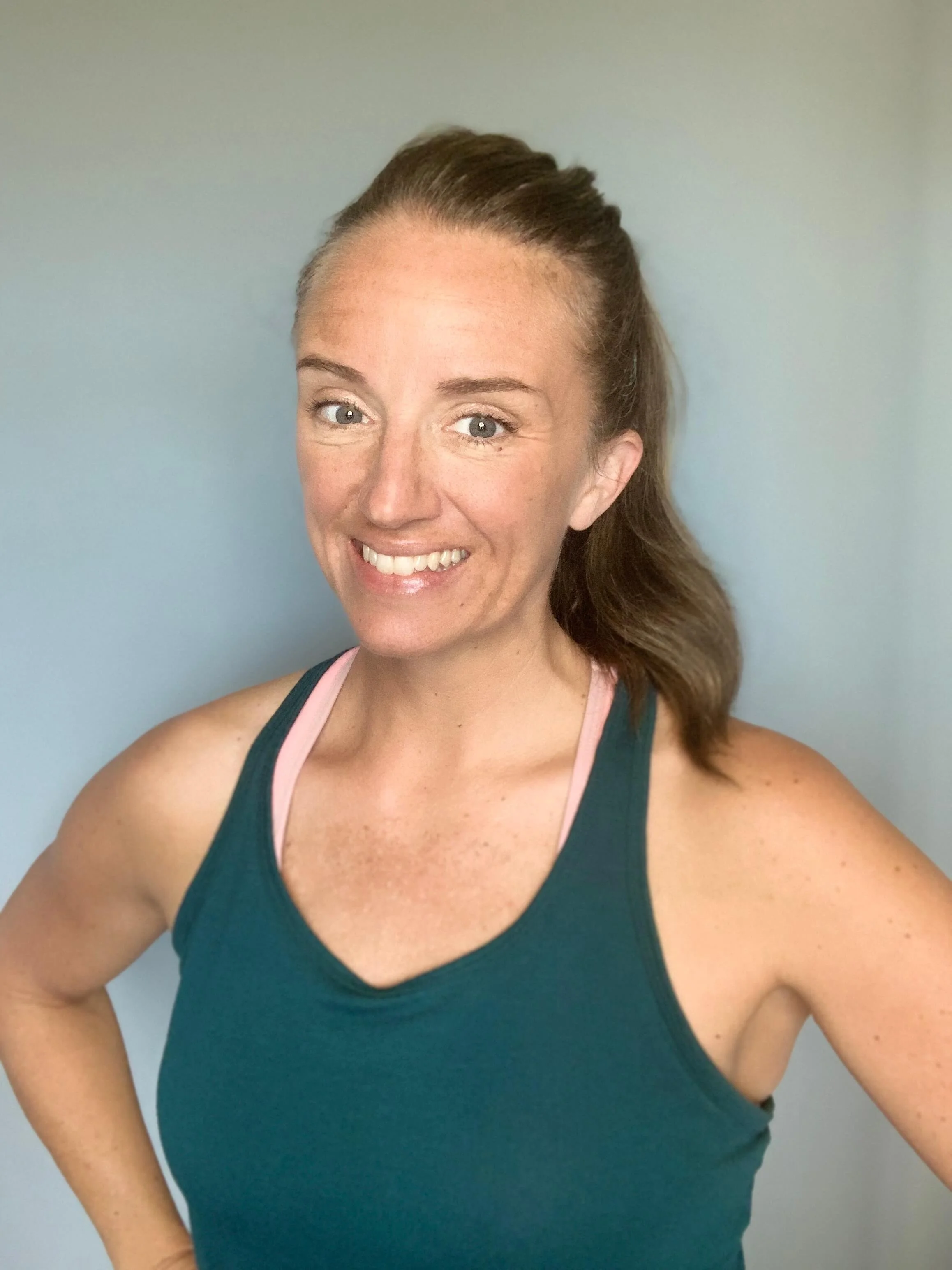 A woman wearing a teal tank top with pink straps, smiling and looking at the camera, with a plain light-colored background.