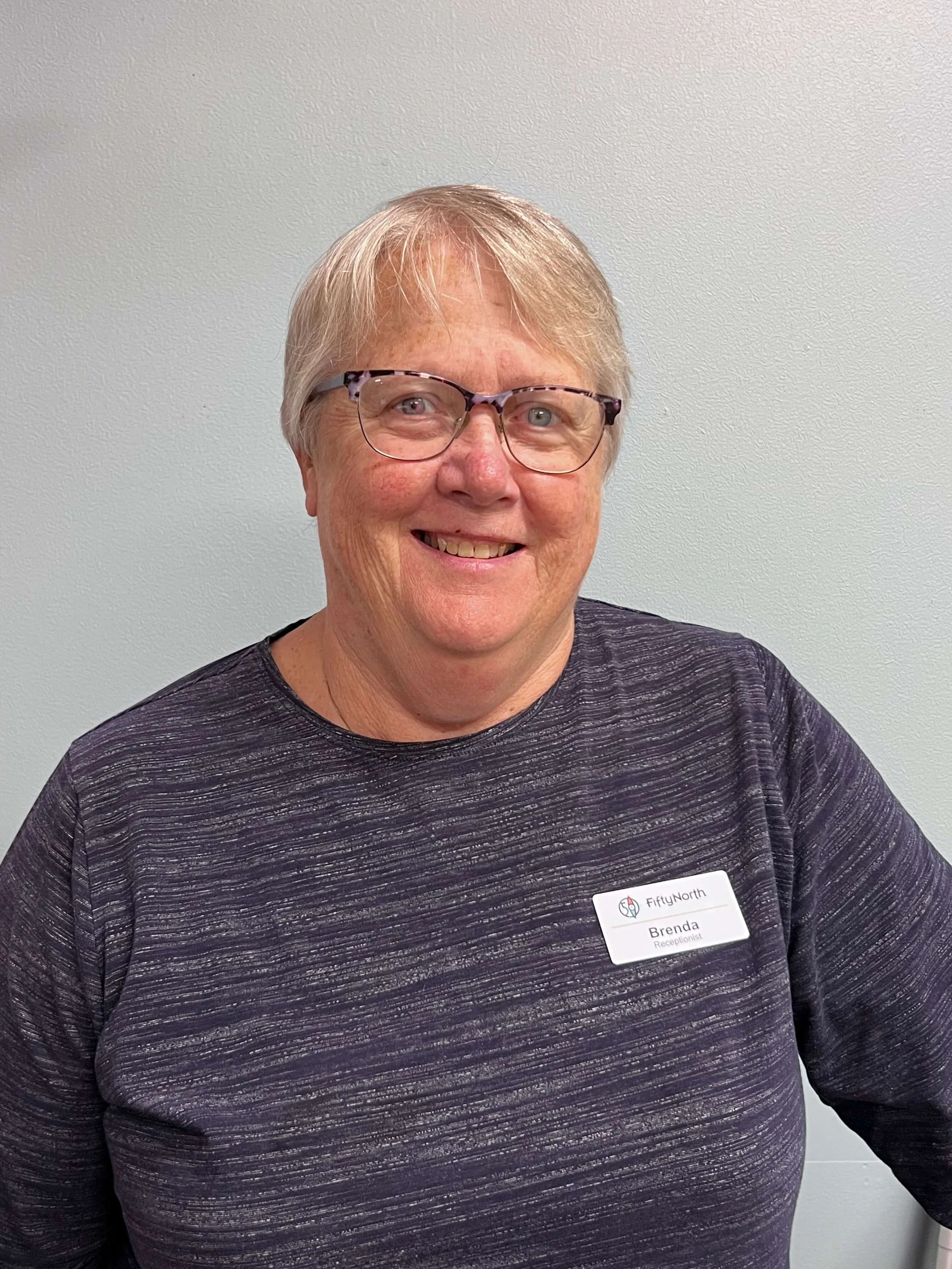 A woman with short blonde hair wearing glasses and a purple patterned shirt, smiling. She has a name tag that reads 'Brenda' and includes 'FiftyNorth' and 'Receptionist'.