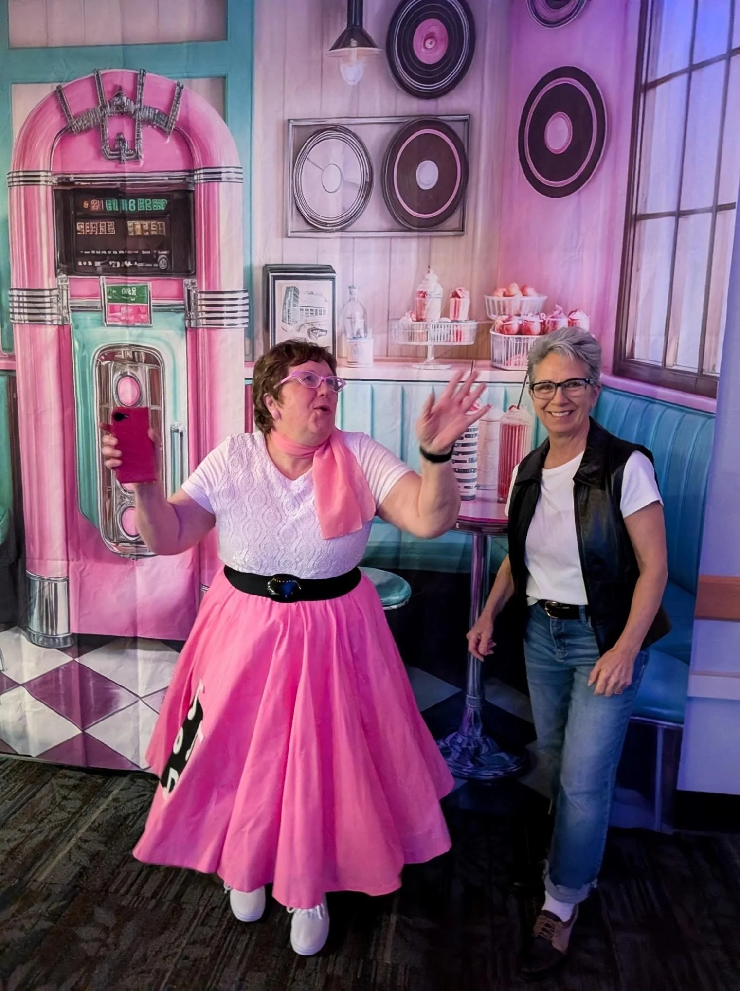 Two women in a colorful retro-themed diner background. One woman is dressed in a pink vintage dress with a black belt and white shoes, holding a pink smartphone. The other woman wears glasses, a white t-shirt, and a black vest. They are smiling and engaging with each other.