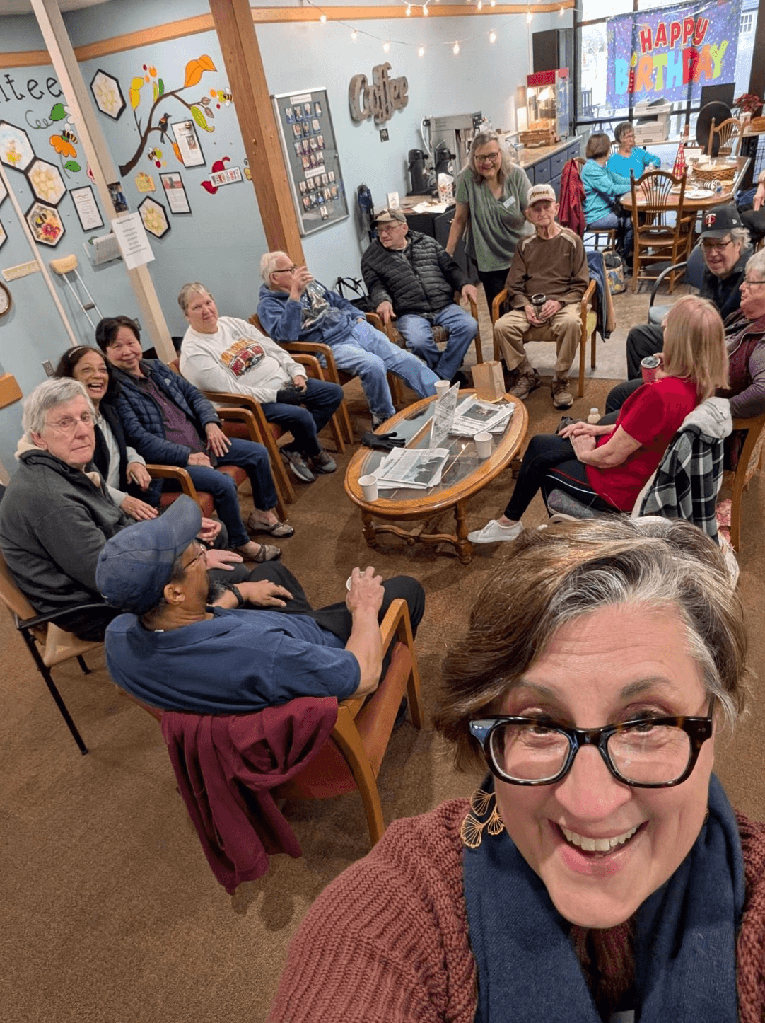 A group of people gathered in a cozy cafe with a 'Happy Birthday' banner in the background. They are sitting on chairs around a coffee table, smiling and engaging in conversation. A woman is taking a selfie in the foreground.