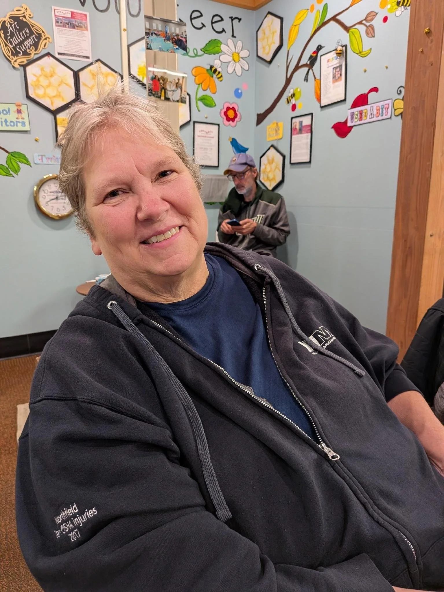 Smiling older woman with short gray hair wearing a dark zip-up jacket, sitting in a room with colorful wall decorations, and a man in the background wearing a cap and using a mobile phone.