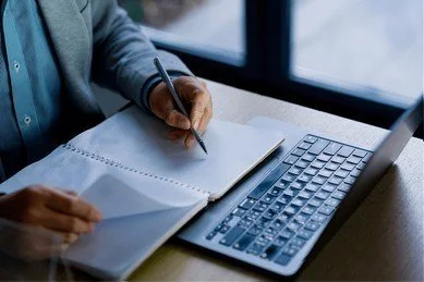 Person writing in a notebook next to a laptop on a desk.