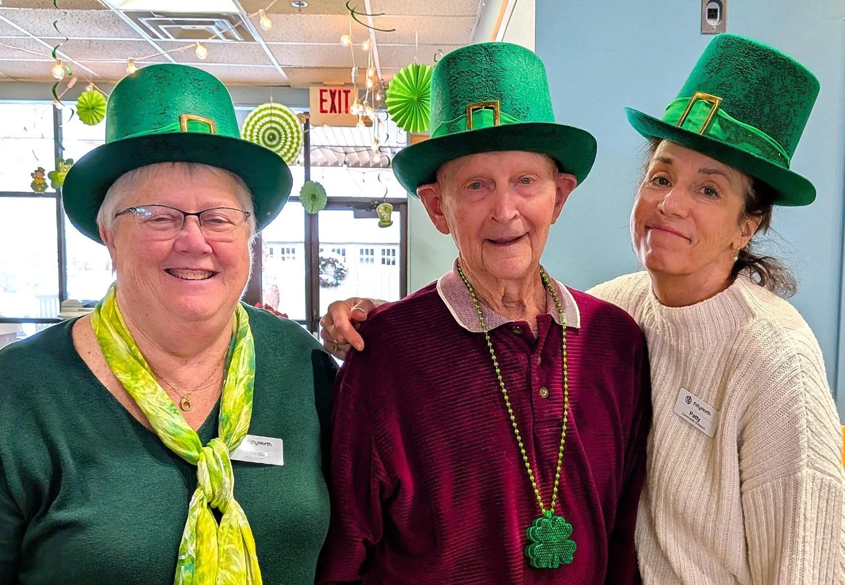 Three women celebrating St. Patrick's Day, wearing large green leprechaun hats, with festive decorations in the background including green paper fan decorations, hanging string lights, and an exit sign.