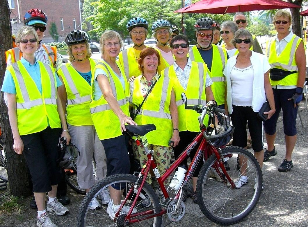 A group of elderly people wearing bright yellow safety vests and bicycle helmets, standing outdoors with a bicycle in front of them. Some are smiling and appear to be enjoying a bike ride or outdoor activity.