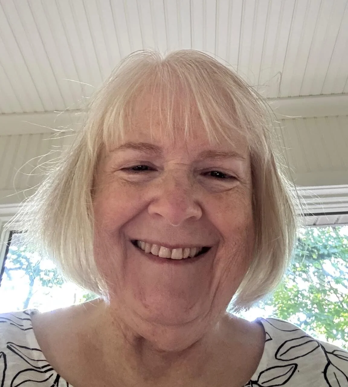 Close-up of a smiling woman with short, light blonde hair, wearing a white top with black leaf patterns, with a background of a window showing green foliage outside.